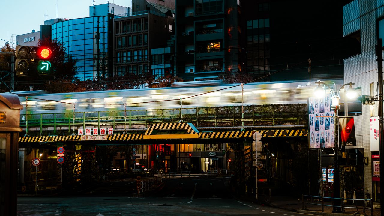 A train blurs across a bridge over a city street at dusk, the traffic light on red.