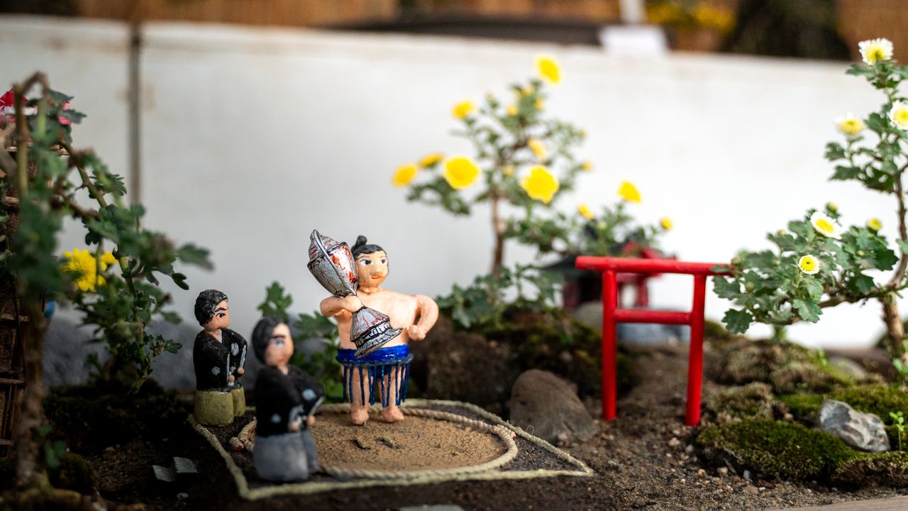 A tiny sumo figure lifting a trophy in a miniature garden with a red torii gate.
