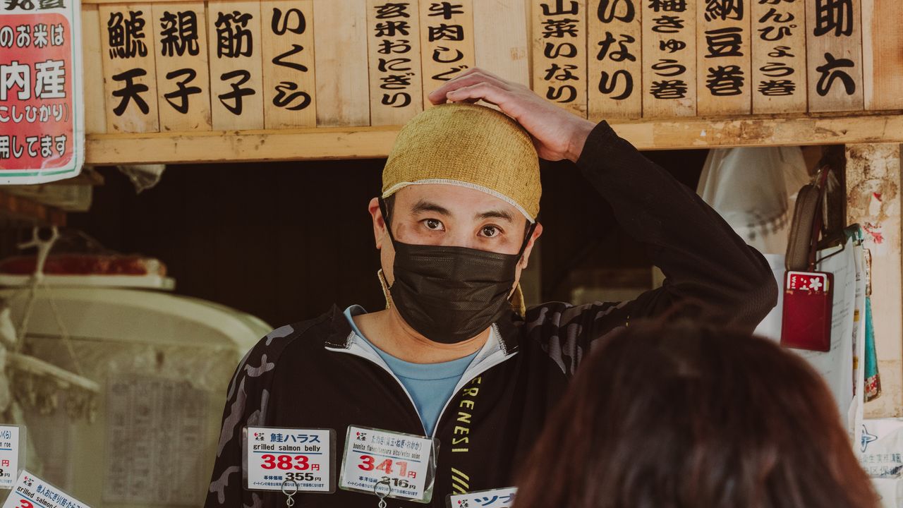 A masked stall vendor scratching his head under hand-painted menu boards.