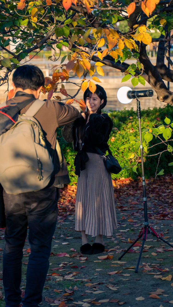 A photographer shooting a smiling model under autumn leaves, a lamp on a stand beside her.