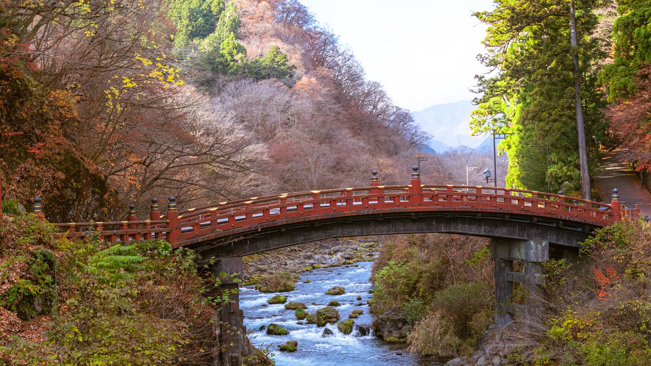 A red arched bridge over a rocky river among autumn trees.