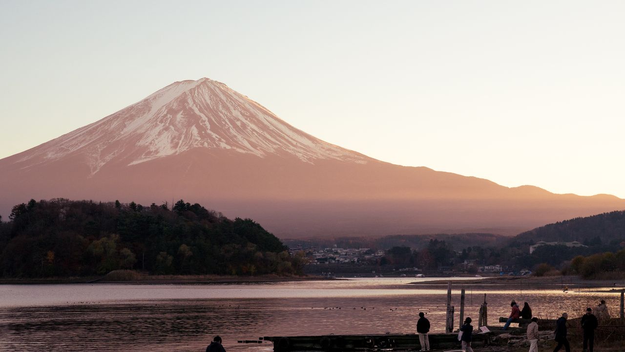 Mount Fuji glowing pink at dusk above a lake, people on the shore.
