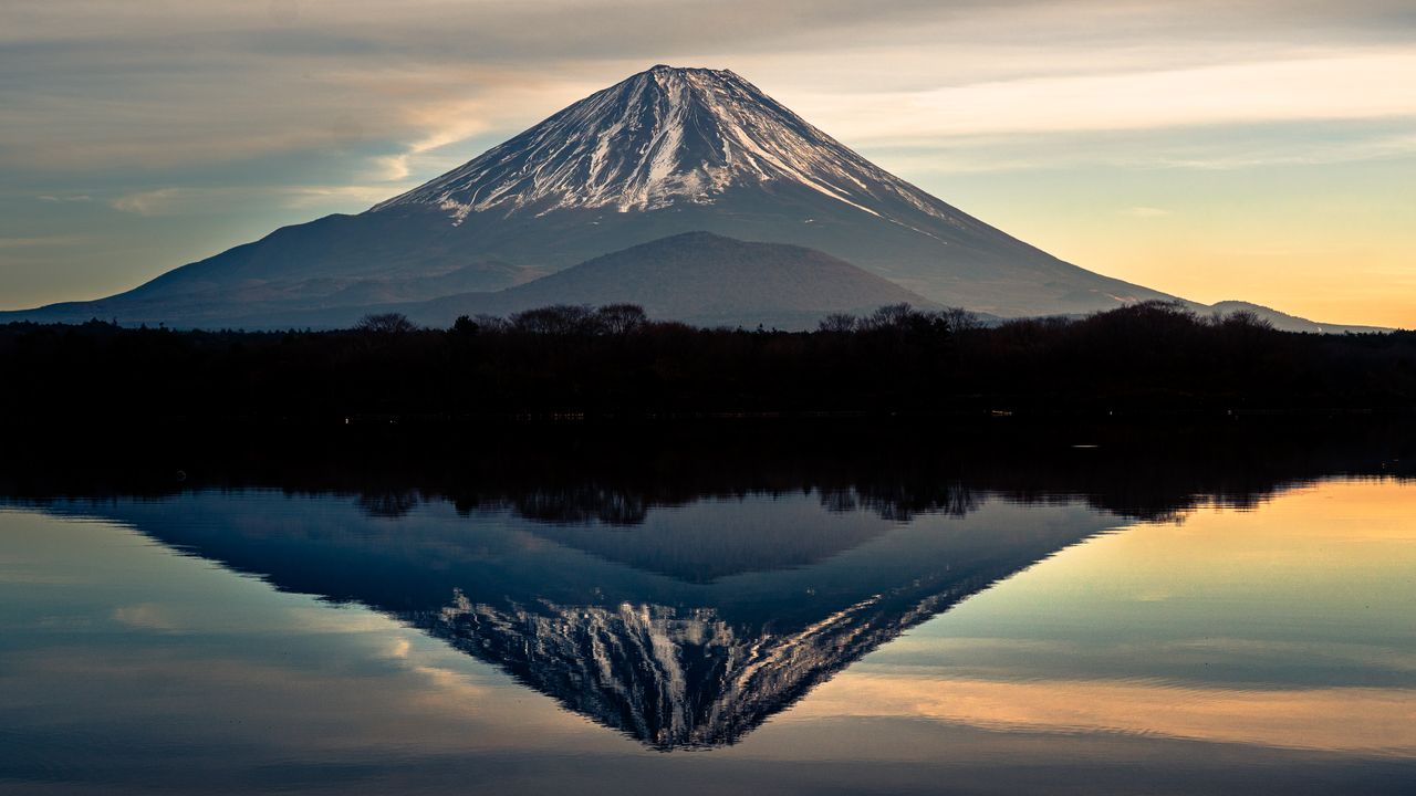 Mount Fuji mirrored in a still lake at dusk, a dark band of forest between.