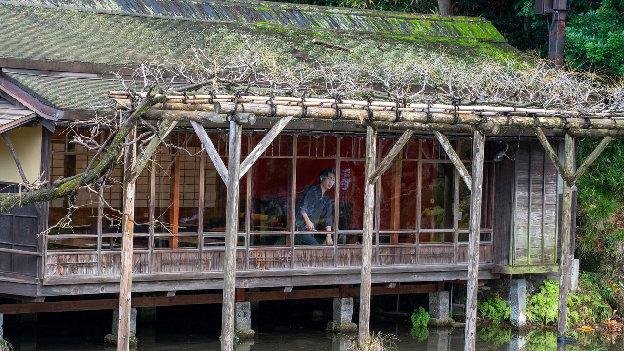 A man inside a wooden teahouse over a pond, seen through a bare wisteria trellis.