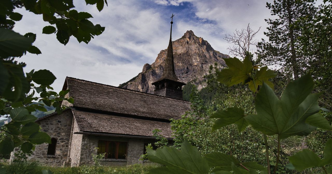 A stone church with a shingled spire below a rocky peak, framed by leaves.