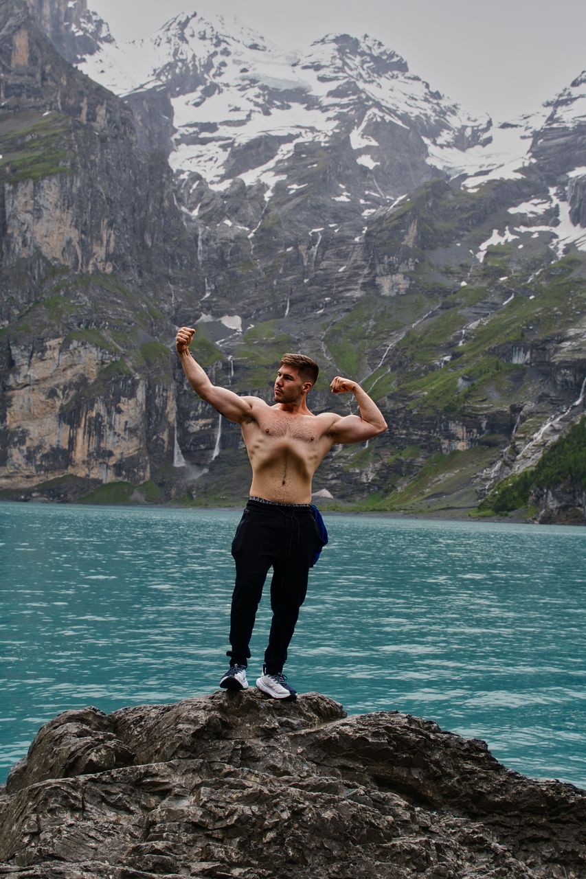 A shirtless man flexing on a rock above a turquoise lake, snowy cliffs behind.
